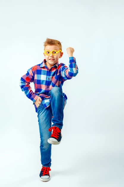 Niño pequeño vistiendo camisa a cuadros de colores, blue jeans, gumshoes, gafas de plástico divirtiéndose en el estudio.