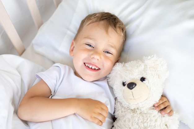 Un niño pequeño yace en una cuna con un oso de peluche en ropa de cama blanca. El sueño de un niño sonríe y mira la cámara.