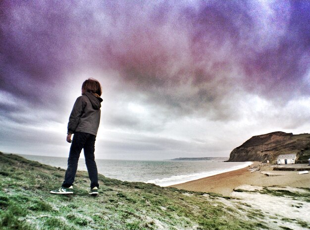 Foto niño de pie en la hierba junto al mar contra el cielo nublado