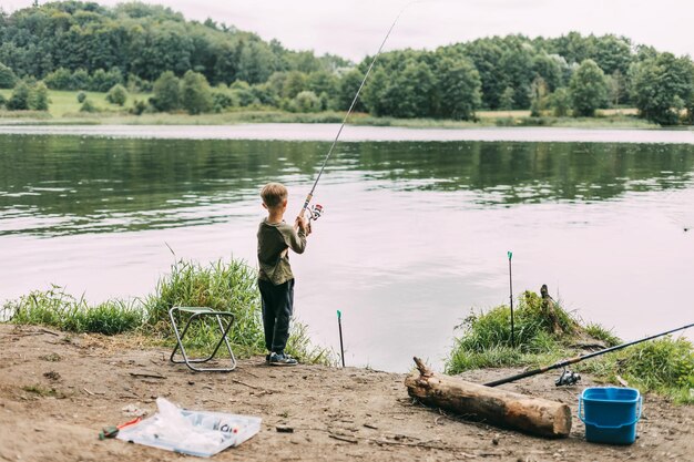 Foto un niño está de pie en la orilla de un lago o río con una caña de pescar durante unas vacaciones familiares