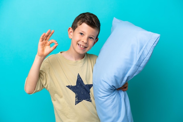 Niño en pijama aislado sobre fondo azul que muestra el signo de ok con los dedos