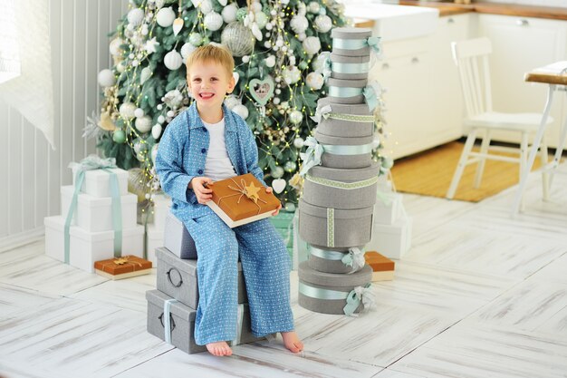 Niño en pijama azul sentado en cajas con regalos de Navidad en el fondo de un árbol de Navidad y un montón de regalos.