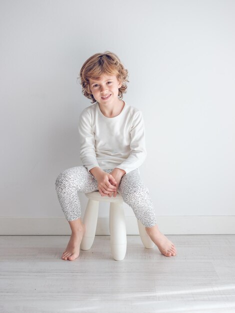 Niño en pijama blanco apoyado contra la pared blanca de su casa.