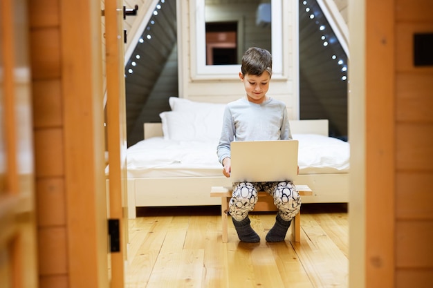 Niño en pijama sentado con una computadora portátil en una cabaña de madera Concepto de felicidad de la actividad de ocio infantil