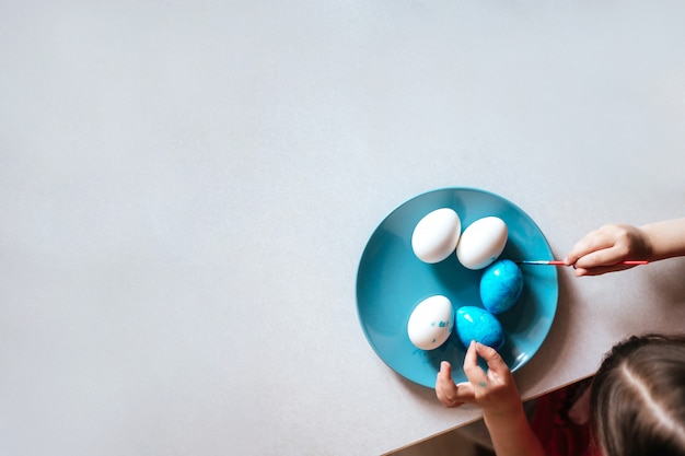 Niño pinta los huevos de Pascua en un plato azul sentado en la mesa de cerca copia espacio