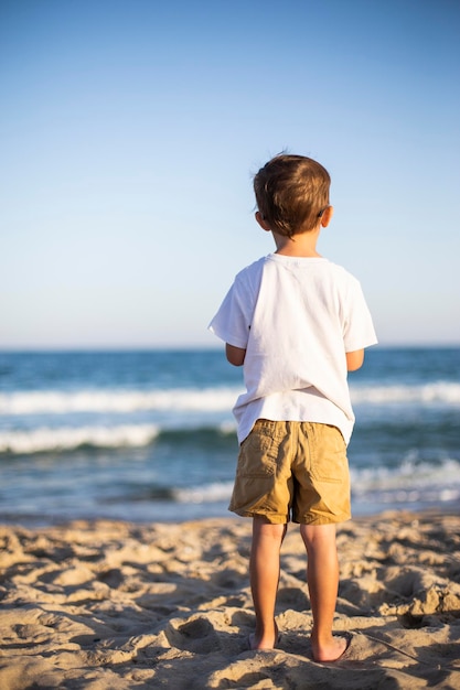 Niño en una playa mirando al mar Niño de pie en la playa mirando al océano