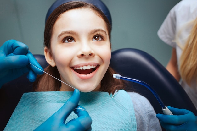 Niño positivo alegre sentarse en la silla dental. Manos en guantes de látex para usar hilo dental en los dientes frontales. Mujer asisstante se encuentra al lado.