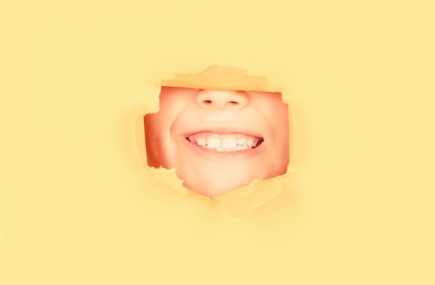 Foto niño positivo con una sonrisa agradable con dientes en la cara se mantiene a través de un agujero rasgado en papel amarillo fondo de papel roto concepto de emociones niño con una sonrisa con dientes muestra la cara en un agujero de papel