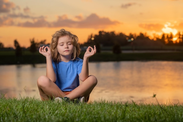 Foto niño practica yoga y medita en posición de loto cerca del lago al atardecer