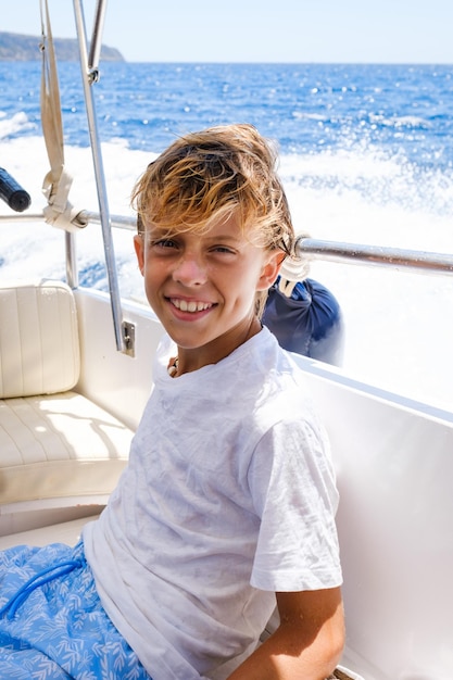 Niño preadolescente alegre con cabello rubio mojado en camiseta blanca sonriendo y mirando a la cámara mientras se sienta en un barco moderno durante un crucero en el mar en un día soleado