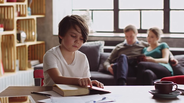 Niño preadolescente leyendo un libro mientras sus padres se relajan en el sofá en segundo plano.