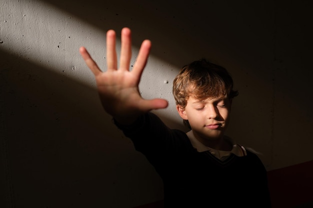 Niño preadolescente pensativo de pie con los ojos cerrados en la pared de piedra demostrando un gesto de parada con la palma de la mano hacia un brillante rayo de sol inesperado desde la ventana