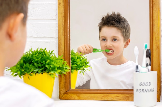 Foto niño preadolescente en pijama cepillándose los dientes con un cepillo de dientes y mirando su reflejo en el espejo del baño por la mañana, vista posterior.