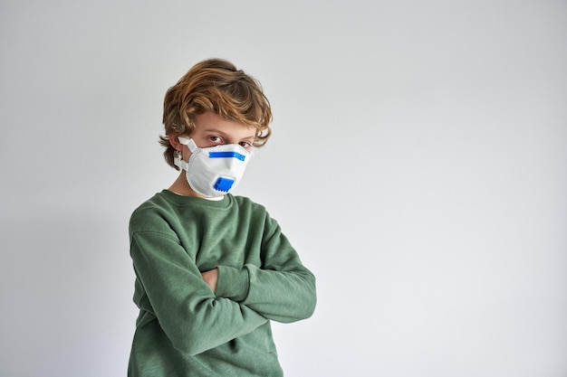 Niño preadolescente en respirador médico con brazos cruzados durante las restricciones de cuarentena durante la pandemia de covid19 y mirando la cámara sobre fondo gris