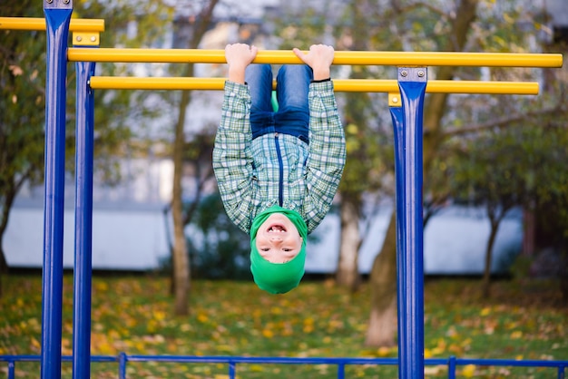 Niño preadolescente con sombrero verde sobre duela de metal al aire libre. Niño de escuela en barra horizontal boca abajo