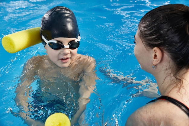 Niño preescolar aprendiendo a nadar en la piscina con fideos de espuma con entrenador joven