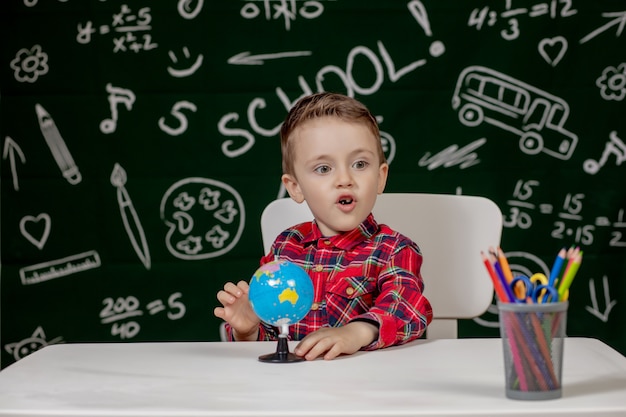 Niño preescolar haciendo la tarea escolar. Escolar con expresión de la cara feliz cerca de escritorio con útiles escolares. Educación. La educación primero. Concepto de escuela