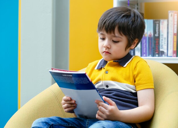 Foto niño preescolar leyendo un libro con cara curiosa en la biblioteca con fondo borroso de estantería