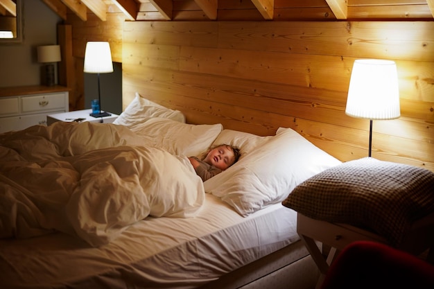 Un niño preescolar que usa pijamas está durmiendo en la cama de sus padres en casa por la noche con la luz encendida