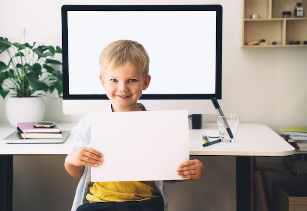 Niño preescolar sosteniendo en las manos papel blanco en blanco sobre el fondo de la computadora con pantalla blanca en blanco en el interior del hogar Niños aprendiendo en línea desarrollo de educación a distancia Educación en el hogar para niños