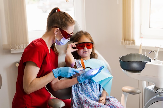El niño se prepara para la iluminación ultravioleta del relleno dental con fotopolímero en odontología El dentista con gafas protectoras rojas trata y elimina la caries en una paciente niña