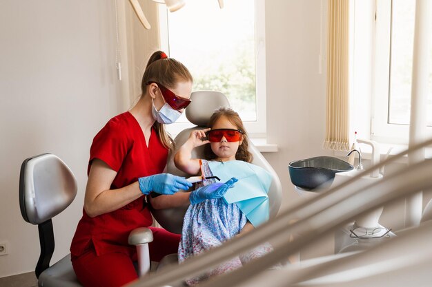 El niño se prepara para la iluminación ultravioleta del relleno dental con fotopolímero en odontología El dentista con gafas protectoras rojas trata y elimina la caries en una paciente niña