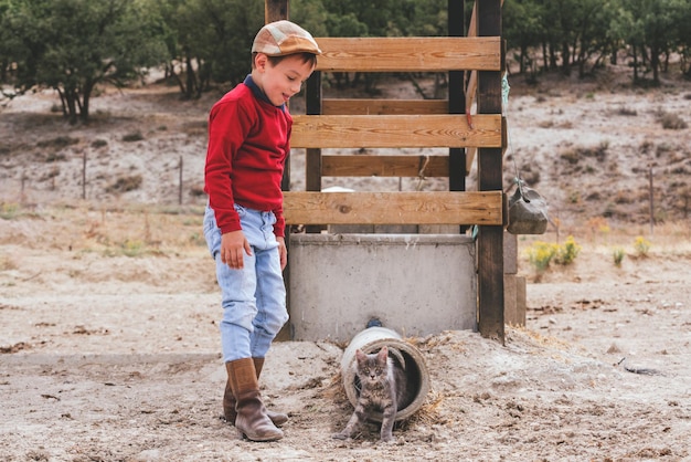 Niño de pueblo caucásico con botas y una boina jugando con un gatito en una granja en un día soleado.