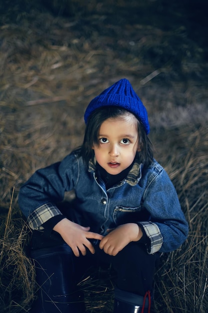 Foto niño del pueblo un niño con botas está sentado en el heno en un granero con ropa cálida y un sombrero azul