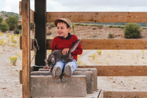 Niño de pueblo sonriente caucásico con botas y una boina sentado con un gatito en una granja en un día soleado.