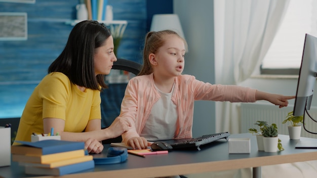 Niño que recibe asistencia para una tarea en línea de la madre en la computadora en casa. Padre ayudando a la niña con la tarea y las lecciones escolares remotas, señalando el monitor. Colegiala y mamá