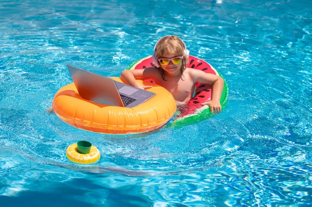 Niño que viaja de verano trabajando en una computadora portátil en la piscina de verano pequeño profesional independiente que usa computadora remota