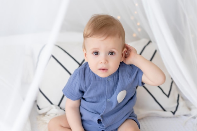 Un niño recién nacido con un traje azul se sienta en una cama con un velo en el interior de un dormitorio en colores pastel. Mira a la cámara los productos de los niños de la mañana de los niños felices.