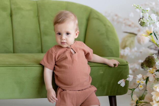 Un niño recién nacido con traje beige se para cerca de un sofá verde rodeado de hermosas flores Mirando la cámara