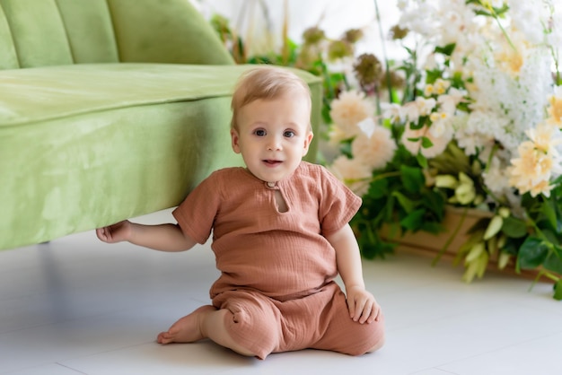 Un niño recién nacido con un traje beige se sienta cerca de un sofá verde rodeado de hermosas flores Sonriendo mirando a la cámara