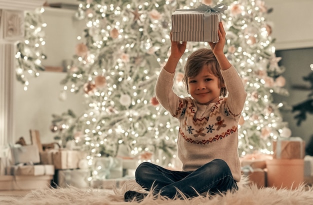 El niño con un regalo sentado en el suelo en el fondo del árbol de Navidad