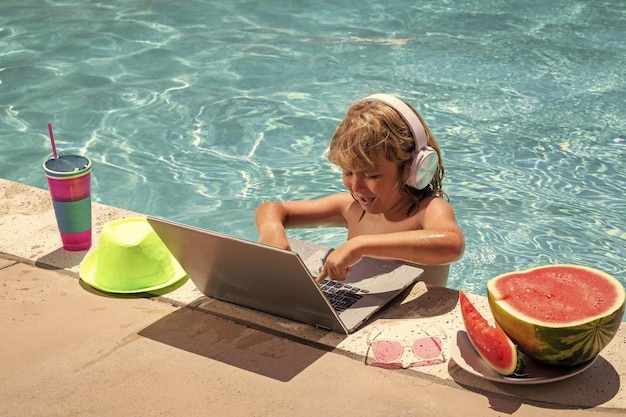 Niño relajándose en la piscina usando una computadora portátil en agua de verano Niño niño estudiando en línea o trabajando en la playa tropical Concepto de tecnología para la vida