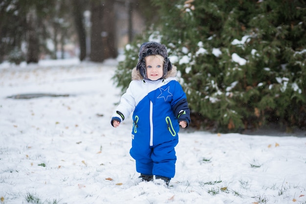 Niño riendo de un año en un cálido traje de nieve y un cazador de gorra caminando en el parque de invierno con una nieve blanca. Primer invierno y primeros pasos del niño pequeño en la nieve