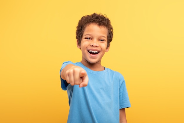 Foto niño riendo apuntando a la cámara con camiseta azul sobre fondo amarillo