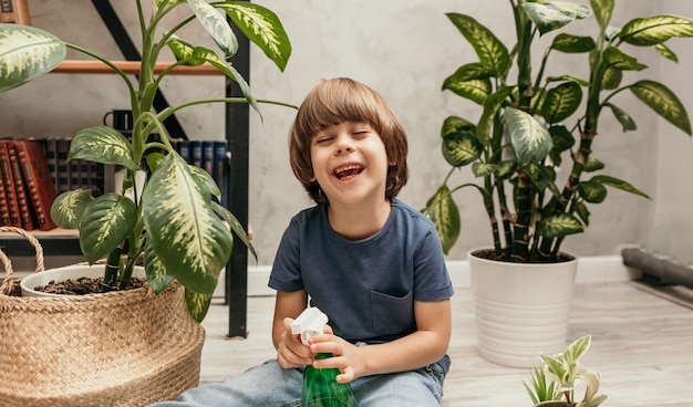 Un niño riendo se sienta en el suelo en una habitación con plantas y sostiene un rociador