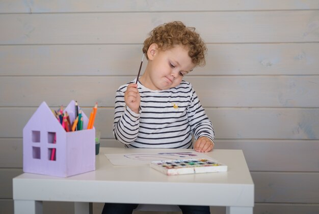 Niño rizado dibuja pinturas de acuarela sobre papel sentado en la mesa