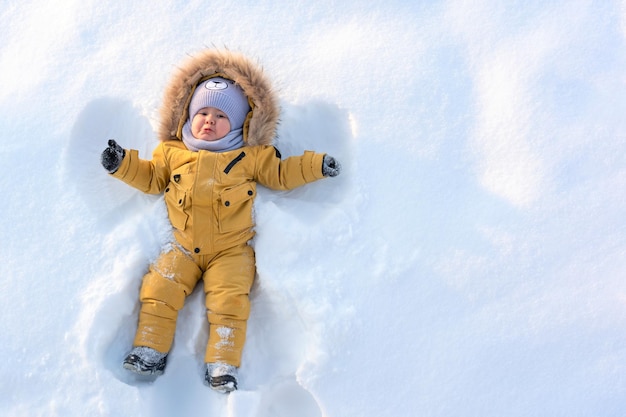 Un niño con ropa amarilla de invierno yace en la nieve y hace un ángel de nieve copiar espacio