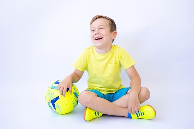 Un niño con ropa deportiva está sentado sobre un fondo blanco sosteniendo una pelota de fútbol en sus manos y tiene un estilo de vida saludable