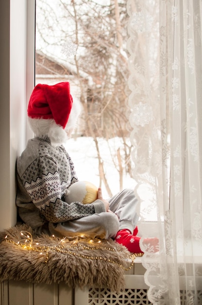 Un niño con ropa festiva y un sombrero rojo de Navidad está sentado junto a la ventana con una bola dorada en la mano.