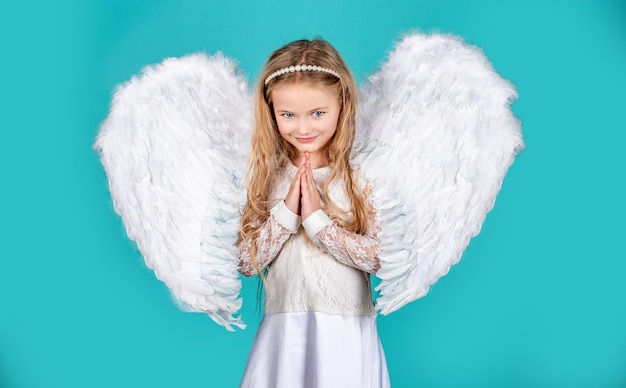 Niño con rostro angelical. Muchacha linda del niño que presenta con alas de ángel. Hermosa niña ángel de pie con los brazos cerca del pecho como en oración. Pequeñas manos de ángeles se cruzaron por ti.