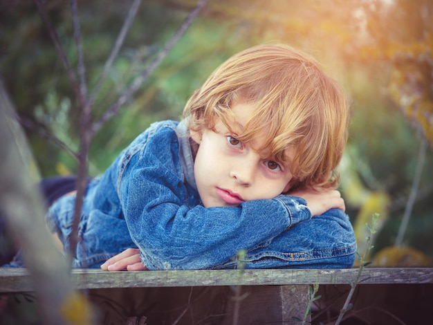 Un niño rubio aburrido con chaqueta vaquera doblando los brazos y mirando a la cámara mientras yacía en el paseo marítimo entre ramitas en el campo