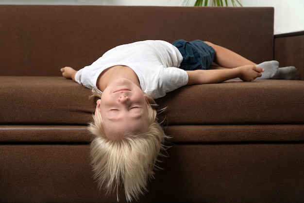 El niño rubio se acuesta en el sofá con la cabeza colgando El niño está cansado Posición incorrecta para dormir del niño