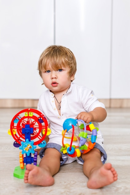 Un niño rubio de apariencia europea está sentado en el piso de la casa y jugando con juguetes de colores brillantes