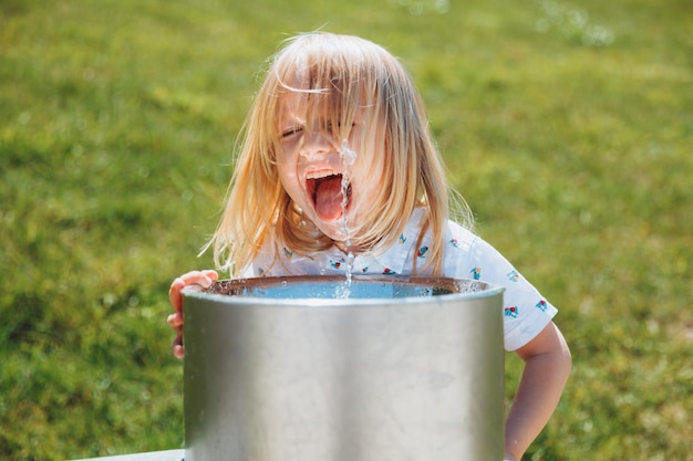 Un niño rubio bebe de una fuente de agua potable en un parque de la ciudad en un caluroso día de verano al aire libre