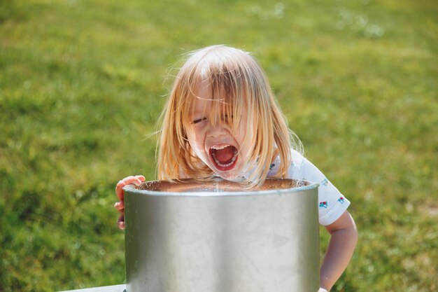 Un niño rubio bebe de una fuente de agua potable en un parque de la ciudad en un caluroso día de verano al aire libre