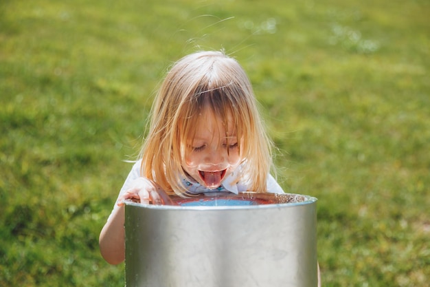 Un niño rubio bebe de una fuente de agua potable en un parque de la ciudad en un caluroso día de verano al aire libre